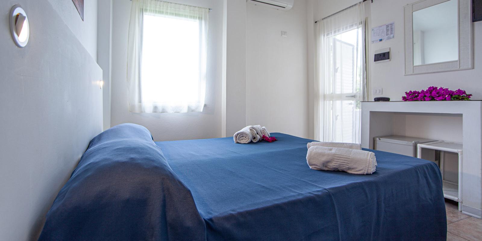 Camera luminosa con letto matrimoniale, lenzuola blu e fiori su un tavolo.
