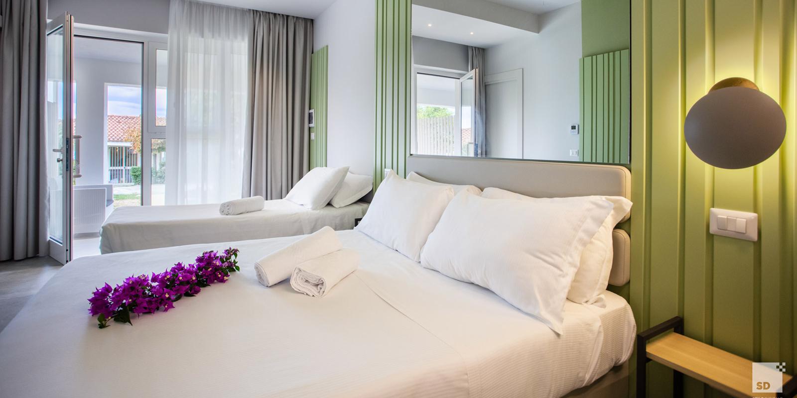 Camera moderna con letto matrimoniale, fiori viola, lenzuola bianche e luce soffusa.
