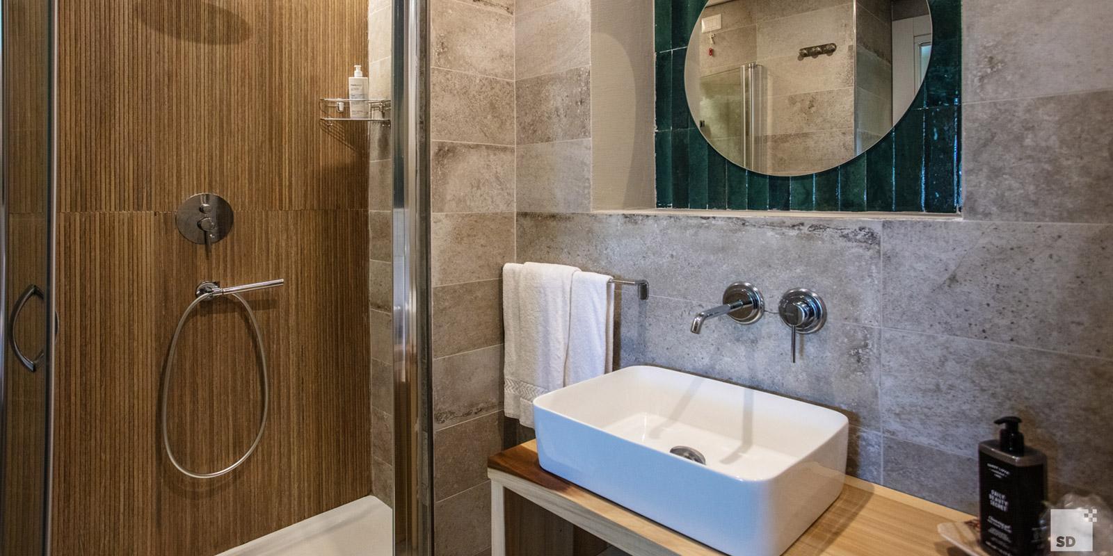 Bagno moderno con doccia, lavabo rettangolare e specchio rotondo su piastrelle verdi.